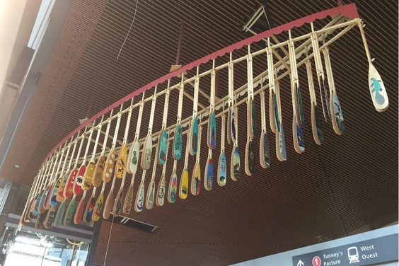 Une structure en forme de canot suspendue au plafond d’une gare, avec des pagaies colorées accrochées à la structure.