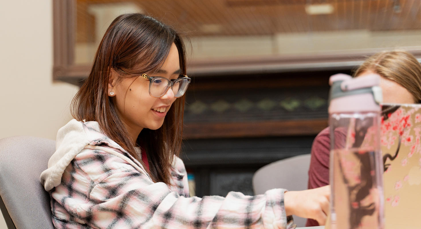 A smiling student wearing glasses works enthusiastically on her laptop in a campus study space.