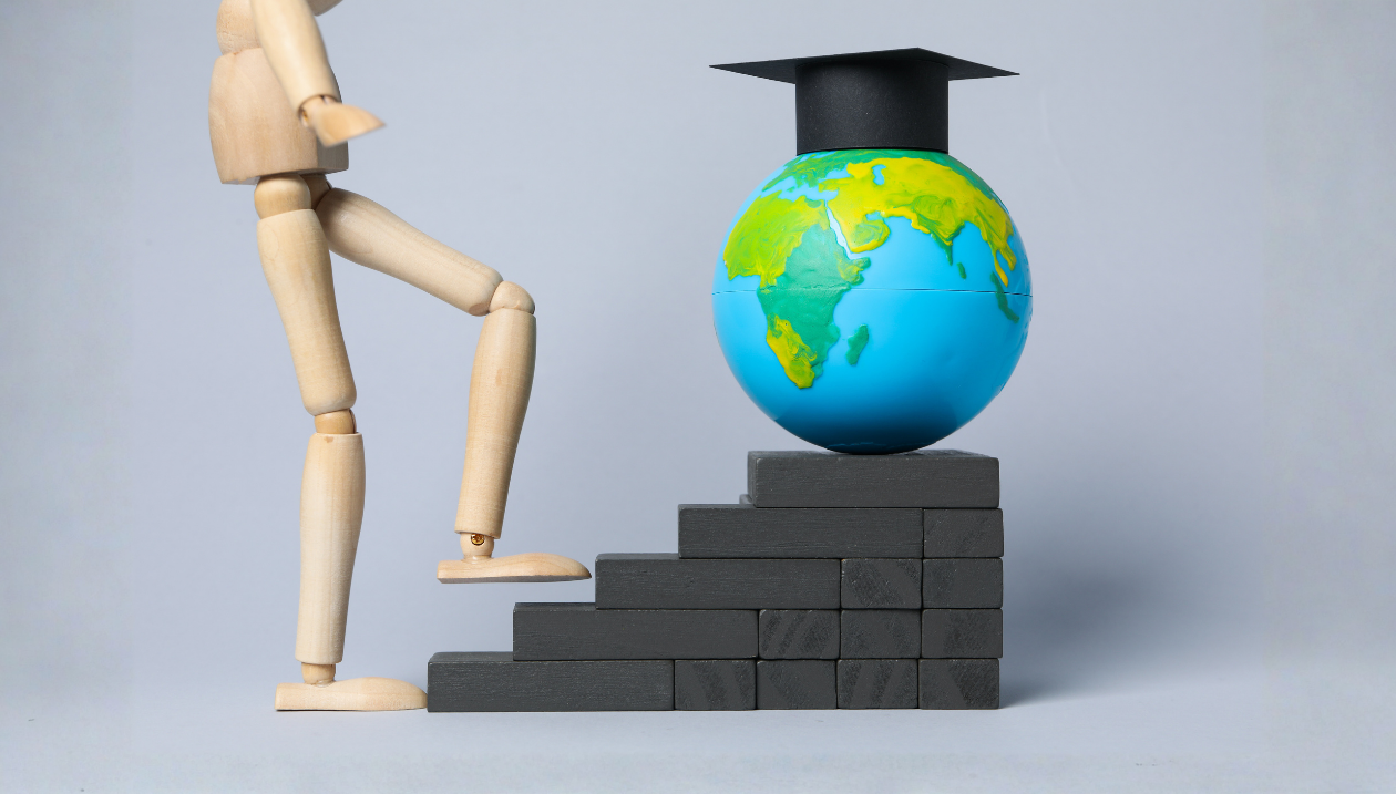 wooden figure about to climb steps toward globe with mortarboard, representing academic journey