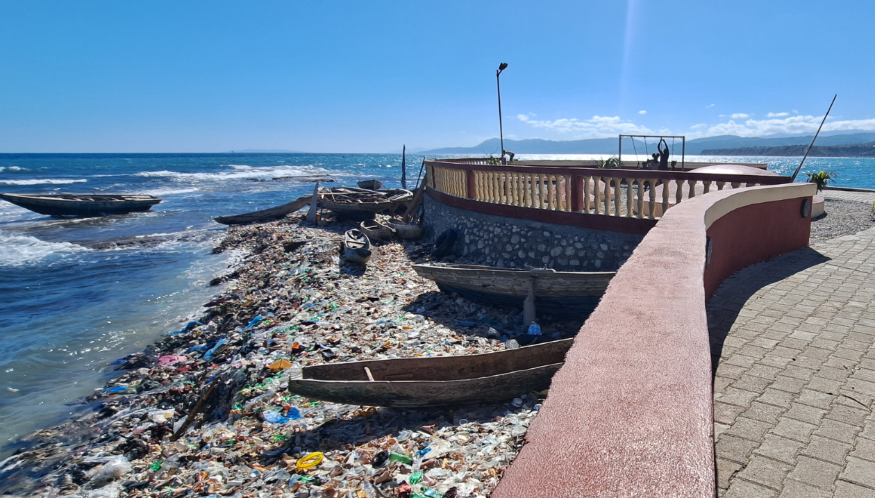 A coastal scene at Fort La Pointe in Jérémie, Haiti. Several wooden fishing boats rest along the shoreline beside a large accumulation of plastic waste and other debris. A renovated waterfront promenade with stone walls overlooks the beach, illustrating the contrast between heritage revitalization, artisanal fishing and ongoing environmental challenges.