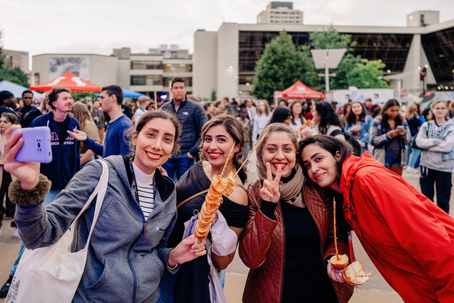 Four students smile for a selfie while enjoying food at a busy outdoor campus event, with a crowd gathered in the background. 