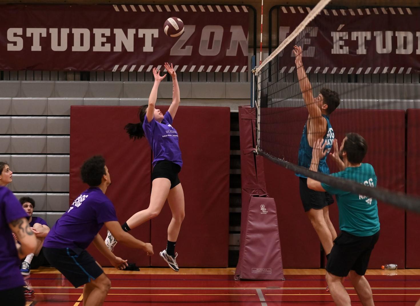 Une joueuse de volley-ball saute pour faire une passe, tandis que les joueurs adverses sautent vers le filet pour contrer.
