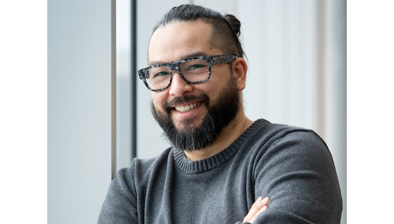 Portrait d’un homme souriant portant des lunettes et une barbe, vêtu d’un chandail gris foncé et les bras croisés devant une fenêtre lumineuse.
