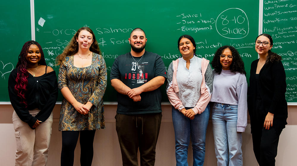 Six étudiantes et étudiants ayant participé en 2025 à un défi d'apprentissage expérientiel interdisciplinaire organisé par la Faculté des sciences sociales se tiennent devant un tableau noir dans une salle de cours.