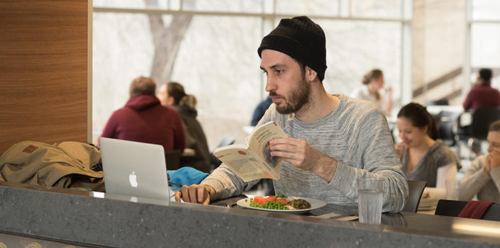 Un homme portant une tuque et tenant un livre utilise son ordinateur portable en mangeant.
