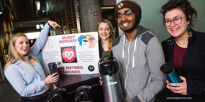 Trois femmes et un homme tiennent des tasses à café réutilisables, une femme point vers une affiche Les matinées caféinées.