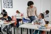 Students writing at their desks in a classroom. A teacher speaks with a student.