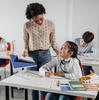 Students writing at their desks in a classroom. A teacher speaks with a student.