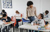 Students writing at their desks in a classroom. A teacher speaks with a student.