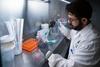 A healthcare researcher in a lab coat works inside a laboratory biosafety cabinet, handling samples with precision tools.