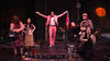 A diverse cast of actors in costume perform on a colorful, dynamic theater set.
