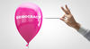 A pink balloon with the word "DEMOCRACY" written on it, being approached by a hand holding a needle, ready to pop it.