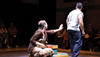 A seated woman reaches toward a departing man on stage as the audience watches.