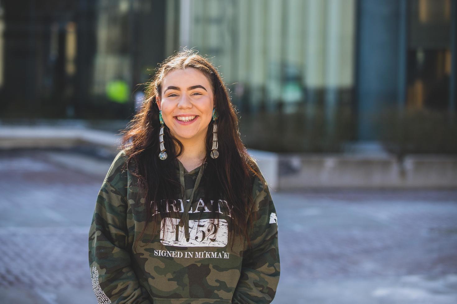 Smiling student with long hair and beaded earrings wearing a green and black sweater in the foreground with a blurred background.