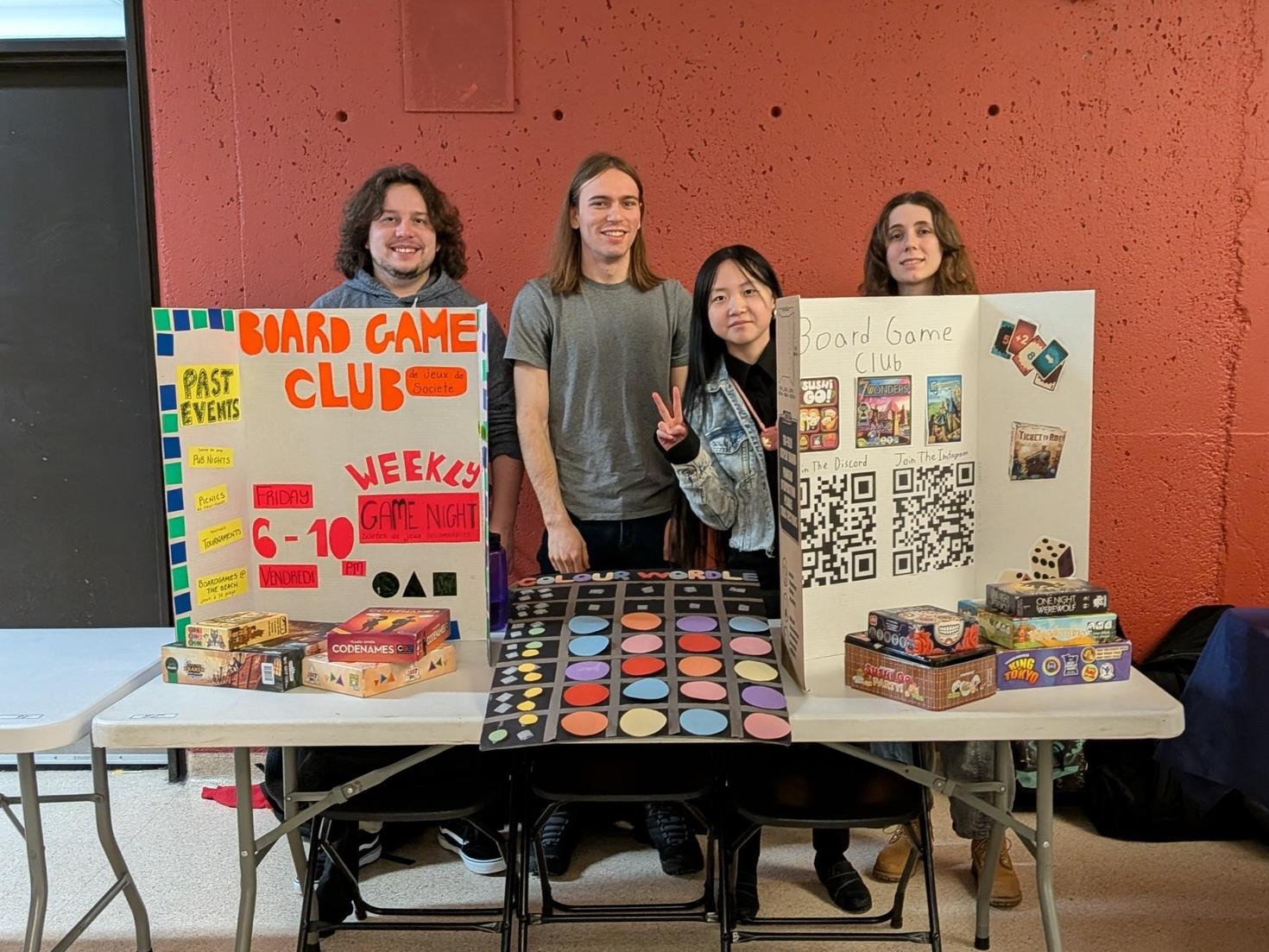 uOttawa Board Game Club executives standing behind their table at the Winter Clubs Fair, with two tri-fold displays, piles of board games, and the club’s custom game, Colour Wordle, in the center.