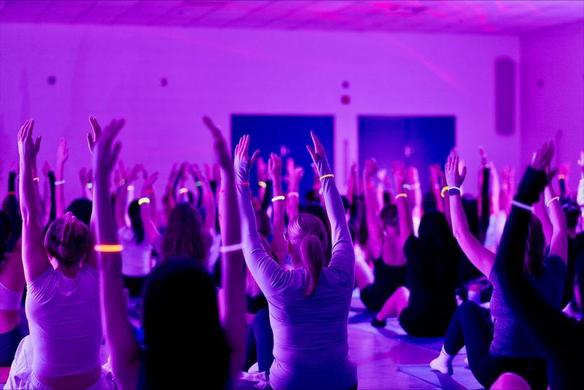 A room filled with students doing Pilates with their arms raised in the air during a Glow-in-the-Dark event.