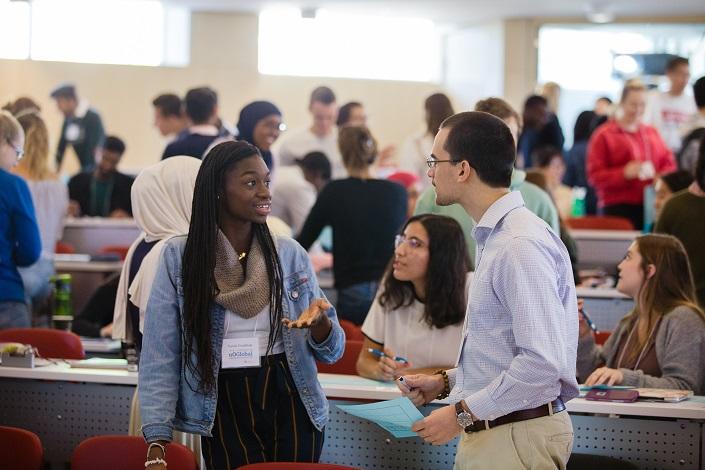 A female and male student chat at the front of a class, with diverse groups of students behind them engaged in their own conversations.