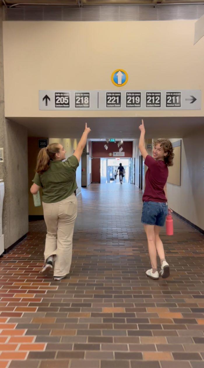 deux étudiantes qui marchent dans un corridor et qui pointent vers un symbole indiquant le chemin raccourci entre deux immeubles