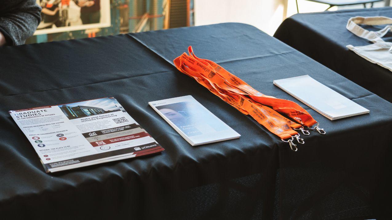 Une vue à plat montre une table recouverte d'une nappe noire, présentant des brochures sur les études supérieures à l'Université d'Ottawa, une plus petite brochure blanche, une pile de cordons orange « uOttawa Engineering » et des brochures blanches.