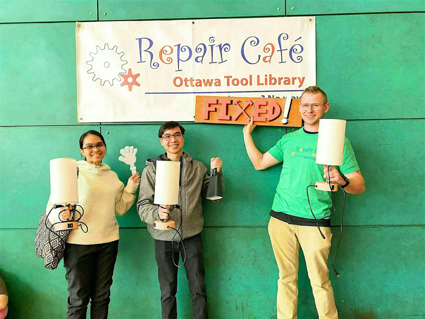 Trois personnes se tiennent devant le logo du Repair Café, avec des lampes et des objets qu’elles ont réparés. 